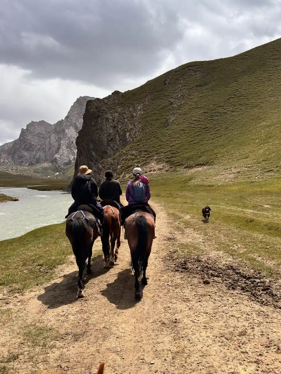 Three riders on horseback with a dog following, riding beside a river along a rocky valley trail
